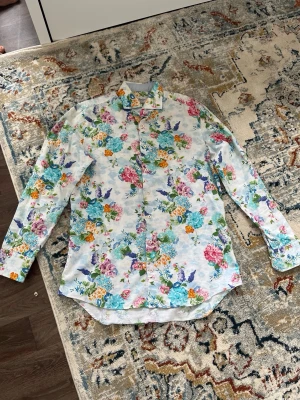 Blommig skjorta med färgglada blommor - Snygg skjorta med färgglatt blommönster i blått, rosa, lila och gult på vit botten. Skjortan har lång ärm, klassisk krage och knäppning framtill. Perfekt för dig som vill sticka ut med en unik stil.