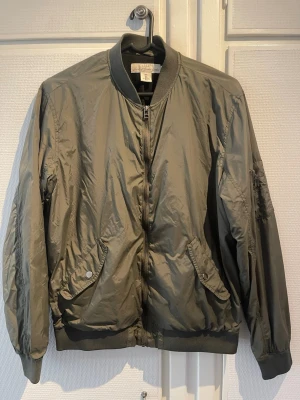 Olivgrön bomberjacka från H&M - Snygg olivgrön bomberjacka från H&M med klassisk ribbad krage, muddar och nederkant. Jackan har dragkedja framtill, två snedställda fickor med tryckknappar och en ärmficka med dragkedja. Tillverkad i glansigt syntetmaterial som ger en cool look.