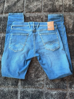 Blå jeans från Replay - Säljer ett par snygga blå jeans från Replay med klassisk femficksdesign och ljusa slitningar. Jeansen har raka ben och detaljerade bakfickor med broderi. Materialet är mjukt denim som ger en skön passform. Perfekt för dig som gillar stilrena och trendiga jeans.