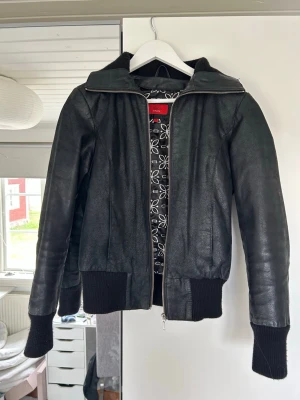 Svart skinnjacka  - Cool svart skinnjacka  med ribbade muddar och krage. Jackan har dragkedja framtill och ett snyggt mönstrat foder med vita blommor. Storlek 38-M men passar bra på 36-S också!