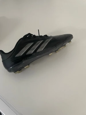 Adidas Copa Pure svarta fotbollsskor - Säljer ett par svarta Adidas Copa Pure fotbollsskor med klassiska silverfärgade stripes på sidan. Skorna har snörning och är tillverkade i syntetmaterial med fasta dobbar för spel på gräs. Perfekta för dig som vill ha stilrena och funktionella skor på planen.