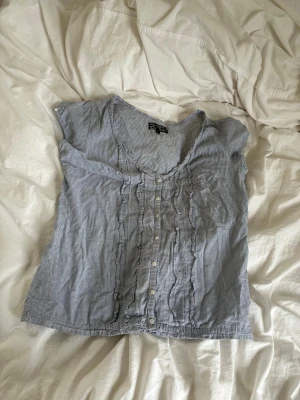 Blårandig blus med volangdetaljer - Söt blårandig blus från H&M med knappar. Blusen har korta ärmar och en lätt v-ringad halsringning som man kan välja hur djup man vill ha den. Tillverkad i ett tunt, mjukt bomullstyg som känns luftigt och bekvämt. Perfekt nu till sommaren och går att både klä upp och klä ner 