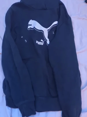 Svart hoodie från Puma med logga - Svart hoodie från Puma med stor vit logga på bröstet. Klassisk modell med huva och känguruficka framtill. Perfekt för en avslappnad och sportig stil. Tillverkad i mjukt material för extra komfort.