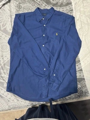 Blå skjorta från Ralph Lauren - Klassisk blå skjorta från Ralph Lauren med gul broderad logga på bröstet. Skjortan har slim fit passform, knappar framtill och lång ärm. Tillverkad i mjuk bomull som känns skön mot huden. Perfekt för en clean och stilren look.
