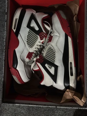 Air Jordan 4 Fire Red sneakers - Säljer ett par klassiska Air Jordan 4 Fire Red sneakers med vit läderöverdel, svarta och röda detaljer samt meshpaneler på sidorna. Skorna har en ikonisk siluett, synlig Air-enhet i sulan och röda accenter på snörningen. Perfekta för dig som gillar streetwear och sneakerskultur. Inga stora tecken på användning 🙏🏻🙏🏻