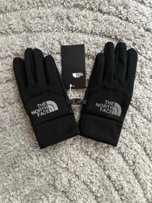 Svarta handskar från The North Face - Svarta handskar från The North Face med logga i vitt på ovansidan. Handskarna har en stilren design och är tillverkade i ett mjukt och smidigt syntetmaterial som passar perfekt för kyliga dagar.