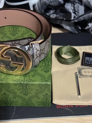 G#CCI Bälte med guldspänne - G#CCI bälte med klassiskt monogrammönster i beige och brunt. Bältet har ett stort, rundat spänne. Tillverkat i skinn och canvas, perfekt för att lyfta vilken outfit som helst.