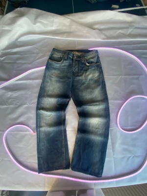 Blå nudie raka jeans med vintage tvätt nudie - Snygga blå jeans med rak passform och klassisk femficksdesign. Jeansen har en vintage tvätt med slitningar och kontrastsömmar. Tillverkade i denim med bälteshällor och knappgylf. Perfekta för en avslappnad och trendig look.41 cm w och 96 ytter och 68 innerben