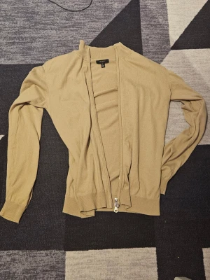 Beige ziptröja från mantinique - Säljer en beige ziptröja från matinique med ribbade muddar och hög krage. Tröjan har dragkedja hela vägen och är tillverkad i ett mjukt, tunt material som passar perfekt till lager på lager. Enkel och stilren design som funkar till det mesta.