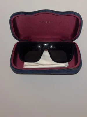 Gucci solglasögon - Snygga svarta solglasögon från Gucci med klassisk fyrkantig form och diskret guldfärgad logga på sidan av bågen. Bågarna är tillverkade i syntetmaterial och kommer med ett fodral i vinröd och grå färg. Perfekt för dig som vill ha en stilren och exklusiv look. Använda fåtal gånger. Skriv för fler frågor🤩