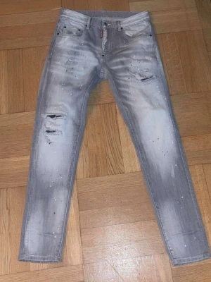 Grå  jeans med färgstänk - Säljer ett par grå  jeans med slitningar och hål på benen samt coola vita färgstänk. Jeansen har fem fickor och en modern, tvättad look som ger en trendig streetstyle-vibe. Perfekta för dig som gillar en edgy stil. Passar dig som är upp till 185 