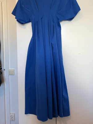 Blå långklänning med v-ringning - En stilren blå långklänning med v-ringad hals och korta ärmar. Klänningen har en luftig passform och faller snyggt längs kroppen. Perfekt för dig som gillar enkel men elegant stil.