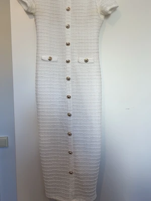 Vit långklänning med knappar - Elegant vit långklänning med korta ärmar och dekorativa knappar längs hela framsidan. Klänningen har två fejkfickor vid midjan och ett diskret strukturerat tyg som ger en exklusiv känsla. Perfekt för dig som gillar klassisk och stilren design.
