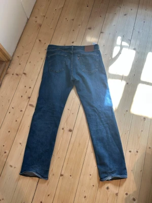 Levi's blå raka jeans - Klassiska blå jeans från Levi's med rak passform. Jeansen har en djupblå tvätt Tillverkade i slitstark denim, perfekta för en avslappnad stil. Lite slitna längst ner vilket visas på följande bild…