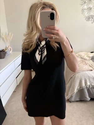 Svart kort stickad klänning - En svart, kort stickad klänning med rund hals och korta ärmar. Klänningen har en enkel och stilren design som passar perfekt till en scarf eller accessoarer. Materialet ser mjukt och bekvämt ut, och modellen har en normal passform.