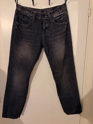 Svarta jeans från Guns Jeans - Säljer ett par svarta jeans från Guns Jeans med klassisk femficksdesign och raka ben. Jeansen har snygga slitningar och kontrastsömmar på bakfickorna. Materialet är denim och passformen är avslappnad. Perfekt för dig som gillar en cool och enkel stil.