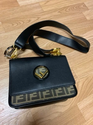 Fendi svart axelväska med FF-mönster - Snygg axelväska från Fendi i svart läder med bred axelrem och guldfärgade detaljer. Väskan har klassiskt FF-mönster i beige och svart på framsidan samt Fendi-logga i metall på locket. Perfekt för dig som gillar lyxiga accessoarer med ikonisk design. +AA// snabbt affär