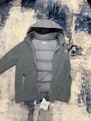 Grå Moncler cardigan -   Säljer en riktigt snygg och bekväm jacka från Moncler. Grå stickad modell med vadderad huva, perfekt för vår/höst eller mild vinter. Väldigt skön på och lätt att matcha till det mesta. Kvitto medföljer och alla tags!  • Dragkedja framtill • Fickor med dragkedja • Moncler-logga på ärmen • Original – tag kvar