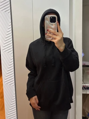 Svart oversized hoodie med ficka - En svart oversized hoodie med stor känguruficka framtill och justerbar huva med dragsko. Hoodien har långa ärmar och ribbade muddar. Perfekt för en avslappnad och cool stil. Materialet känns mjukt och bekvämt, troligen bomullsblandning.