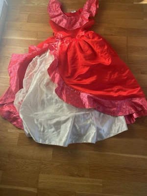 Disney prinsessklänning röd/rosa - Maskeradklänning från Disney i rött och rosa med glittriga mönster och vit underskjol. Perfekt för cosplay eller maskerad, inspirerad av klassiska prinsessor. Klänningen har volanger och glänsande tyg för en sagolik look.