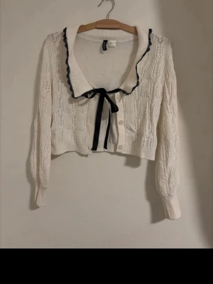 Vit kofta med svart rosett och spets - Söt vit stickad kofta från H&M med svart rosett och svart spetskant vid kragen. Kortare modell med långa ärmar och dekorativa knappar framtill. 