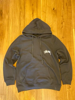 Svart hoodie från Stüssy med tryck - Svart hoodie från Stüssy med klassisk logga på bröstet och stort färgglatt blommotiv samt logga på ryggen. Hoodien har huva med dragsko och en stor magficka. Perfekt för dig som gillar streetwear och vill sticka ut med snygga detaljer.