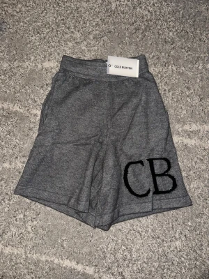 Grå stickade shorts från Cole Buxton - Säljer ett par grå stickade shorts från Cole Buxton med hög midja och breda ben. Shortsen har två bakfickor och ett stort CB-logo broderat framtill. Perfekta för en chill och avslappnad look.