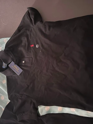 Svart pikétröja från Polo Ralph Lauren - Klassisk svart pikétröja från Polo Ralph Lauren med röd broderad logga på bröstet. Tröjan har krage, knappar vid halsen och korta ärmar. Perfekt för en stilren och avslappnad look.