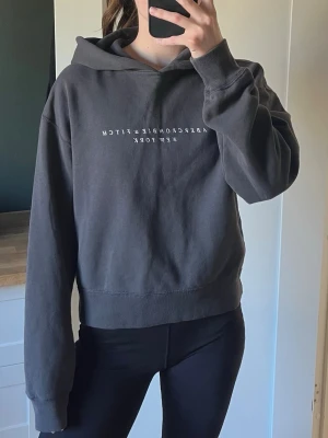 Mörkgrå hoodie Abercrombie & Fitch - Snygg mörkgrå hoodie från Abercrombie & Fitch med vit text 'Abercrombie & Fitch New York' på bröstet. Klassisk passform med huva och ribbade muddar. Perfekt till chill dagar och enkel att matcha med jeans eller tights. Mjuk bomullskänsla.