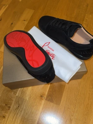 Svarta Louboutin Skor - 43 - Snygga svarta sneakers från Christian Louboutin i mocka med klassisk röd sula och svarta skosnören. Låg modell med diskret logga och stilren design som sticker ut med den ikoniska röda undersidan. Skorna kommer med paket, certifikat och även den originella dustbag.