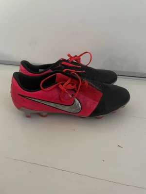 Nike Phantom VNM rosa/svarta fotbollsskor - Säljer ett par Nike Phantom VNM fotbollsskor i en snygg kombination av rosa och svart med silverfärgad swoosh. Skorna har plastdobbar och är tillverkade i syntetmaterial med snörning. Perfekta för spel på gräsplaner och för dig som vill sticka ut på planen.
