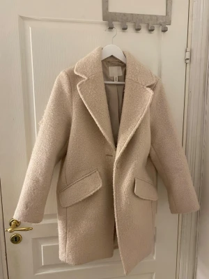 Beige ullkappa från H&M - Säljer en stilren ny och oanvänd beige kappa från H&M med två stora fickor framtill. Kappan är gjord i ett ull-liknande material som känns mjukt och varmt. Perfekt för dig som vill ha en klassisk look med lite oversized passform. Passar S & M i storlek 