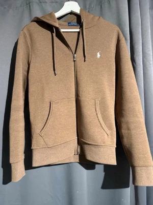 Brun hoodie från Polo Ralph Lauren - Snygg brun hoodie med dragkedja från Polo Ralph Lauren. Klassisk design med huva, dragsnören och två stora fickor framtill. Liten vit logga broderad på bröstet. Perfekt för en avslappnad och stilren look.