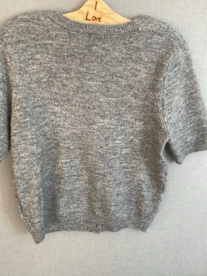 Grå kortärmad kofta med pärlknappar - Grå stickad kofta med korta ärmar och rund hals. Stängs med snygga pärlknappar framtill. Mjuk och skön, perfekt att slänga på sig över en topp. Ribbstickade muddar vid ärmslut och nederkant.