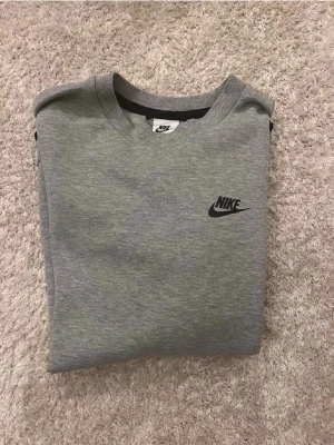 Grå nike tech sweatshirt  - Snygg grå Nike tech sweatshirt med svart logga på bröstet och en cool ficka med dragkedja på ena ärmen. Tröjan har rund halsringning och svarta detaljer vid ärmslut och nederkant. Perfekt till en avslappnad streetwear-look.