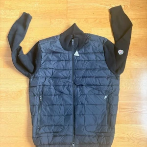 Svart Moncler cardigan - Snygg svart pufferjacka från Moncler med quiltad front och rygg i mjukt material. Jackan har dragkedja framtill, två sidofickor med dragkedja och en Moncler-logga på ärmen. Perfekt för kalla vinterdagar och riktigt stilren look.