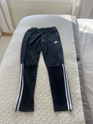 Svarta Adidas träningsbyxor - Säljer ett par svarta träningsbyxor från Adidas med klassiska vita ränder längs benen och Adidas-logga på låret. Byxorna har resår i midjan och är tillverkade i ett lätt och stretchigt material som passar perfekt för sport och träning.