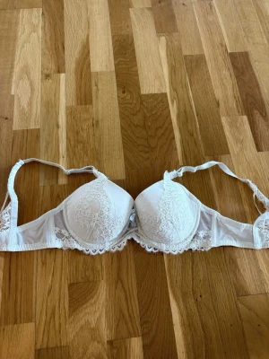 Vit spets-bh med vadderade kupor - Snygg vit bh med vadderade kupor och fina spetsdetaljer framtill och längs kanterna. Axelbanden är justerbara och bh:n har bygel för extra stöd. Perfekt för dig som gillar en feminin och klassisk stil.