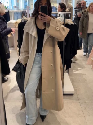 Beige lång trenchcoat med bred krage - Stilren beige trenchcoat med dubbelknäppning, bred krage och justerbara ärmslut. Lång modell som ger en snygg siluett och klassisk look. Perfekt att slänga över en outfit för extra edge. Materialet ser ut att vara bomullsblandning.