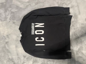 Svart ICON hoodie från Dsquared2 - Svart hoodie från Dsquared2 med stor vit text 'ICON' och 'DSQUARED2' på framsidan. Klassisk passform med ribbade muddar och rund hals. Materialet är mjukt och bekvämt, perfekt för en avslappnad look. Märkt med 'Made in Italy' på etiketten.