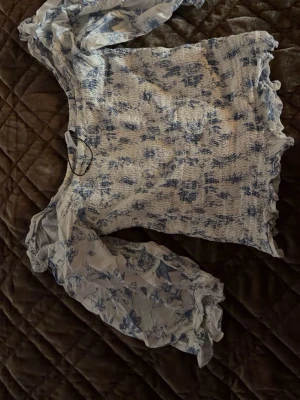 Blommig smockad blus med puffärm - Säljer en vit och blå blus med smockad kropp och blommigt mönster. Blusen har långa, transparenta puffärmar med elastiska muddar och volangdetaljer vid nederkant och ärmslut. Materialet är lätt och luftig, perfekt för en trendig look. Jätte fin och helt oanvänd, köptes i somras. Strl M🤗