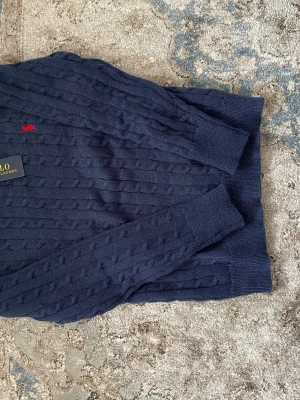 Mörkblå kabelstickad tröja Polo Ralph Lauren - Mörkblå kabelstickad tröja från Polo Ralph Lauren med klassisk röd broderad logga på bröstet. Tröjan har ribbade muddar vid ärmslut och nederkant samt rund hals. Perfekt för dig som gillar tidlös och stilren design.