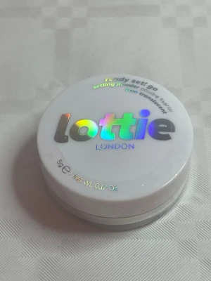 Lottie London Ready Set Go puder - Translucent setting powder från Lottie London i en rund, vit burk med holografisk text. Pudret är veganskt och PETA-godkänt, perfekt för att fixera din bas och ge en matt finish. Innehåller 5g och är lätt att ta med i väskan.