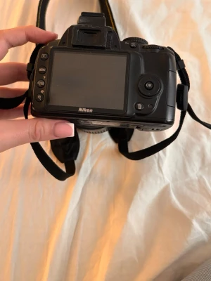 Nikon D3000 systemkamera med objektiv - Säljer en svart Nikon D3000 systemkamera med tillhörande 18-55mm objektiv. Kameran har en stor LCD-skärm, tydliga knappar och en bekväm rem med Nikon-logga. Perfekt för dig som vill ta snygga bilder med en klassisk och pålitlig kamera.