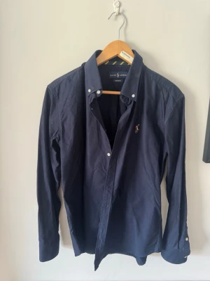Mörkblå skjorta från Ralph Lauren - Klassisk mörkblå skjorta från Ralph Lauren med knappkrage och broderad logga på bröstet. Skjortan har långa ärmar och knappar vid manschetterna. Tillverkad i mjuk bomull för en bekväm känsla och snygg passform. Storlek L men är som S/M