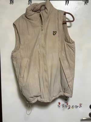 Beige dunväst från Lyle & Scott - Stilren beige dunväst från Lyle & Scott med dragkedja framtill och hög krage. Västen har det klassiska gula fågel-loggan på bröstet och justerbar dragsko nedtill. Perfekt för lager-på-lager och enkel att matcha med olika outfits.