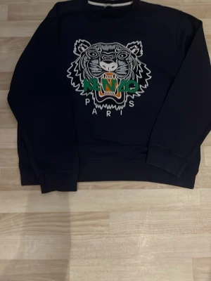 Svart Kenzo sweatshirt med tigertryck - Cool svart sweatshirt från Kenzo med det ikoniska tigerhuvudet broderat framtill i vitt, grönt och orange. Tröjan har rund halsringning och ribbade muddar vid ärmslut och nederkant. Perfekt för dig som gillar streetwear och vill sticka ut.