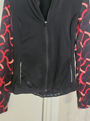 Svart träningsjacka med mönstrade ärmar - Snygg svart träningsjacka med dragkedja framtill och långa ärmar i rött och rosa grafiskt mönster. Tillverkad i ett stretchigt funktionsmaterial som passar perfekt för löpning eller gym. Två fickor framtill och reflekterande detaljer.