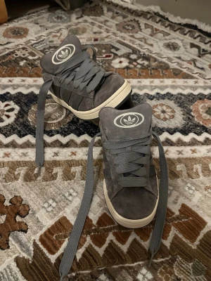 Grå Adidas sneakers i mocka - Snygga grå sneakers från Adidas campus  med klassiska tre ränder på sidan och stort logomärke på plösen. Skorna är i mocka med gråa skosnören och vit sula. Perfekta för dig som gillar streetstyle och vill ha en stilren look. Storlek kan vara från 40-41