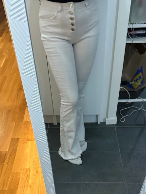 Vita bootcut jeans med knappar - Säljer ett par vita bootcut jeans med hög midja och snygga guldfärgade knappar framtill. Jeansen har råa kanter nertill och sitter tajt över höfterna innan de släpper ut vid benen. Perfekta för en trendig look!